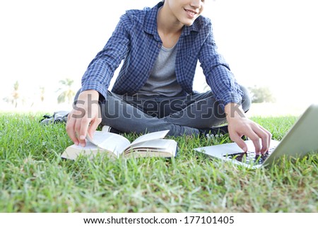 Part view of a teenager boy sitting down on green grass in a park with an open reference book using a laptop to research on-line, studying and smiling. Student outdoors lifestyle.