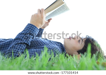 Close up profile portrait of a student teenager boy laying down and relaxing on green grass in a park, reading a text book and studying against a sunny sky. Outdoors lifestyle.