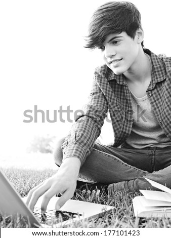 Black and white portrait of a teenager boy sitting on grass in a park with a text reference book and typing on a laptop computer while studying against a sunny sky. Outdoors lifestyle.