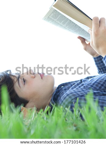 Close up profile portrait of a student teenager boy laying down and relaxing on green grass in a park, reading a text book and studying against a sunny sky. Outdoors lifestyle.