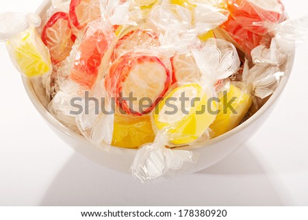 Still life detail view of various candy sweets with orange and lemon fruit flavors wrapped in clear plastic in a kitchen bowl on a white table. Colorful sweet tooth sugar sweets.