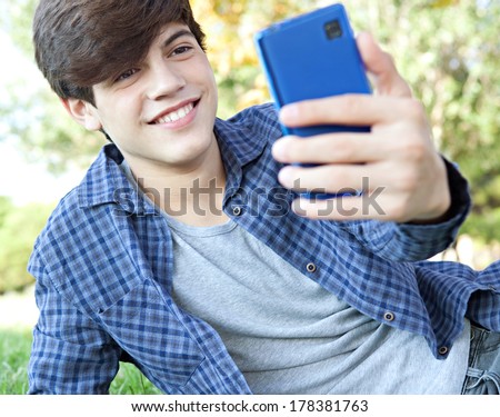 Portrait view of a teenager student boy laying down in a park with his college books, using a smartphone to take a selfie photo of himself for networking, posing and smiling. Technology lifestyle.