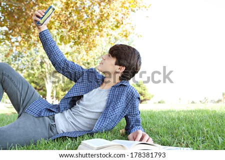Side portrait view of a young teenager student boy laying down in a park with his college books, using a smartphone to take a selfie photo of himself, posing and smiling. Technology lifestyle.