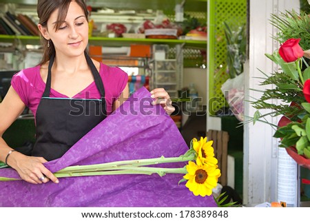Attractive florist business owner woman working at the counter of her flower market store wrapping yellow sunflowers in wrapping paper during a sunny day. Small business owner.