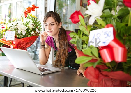 Side portrait of an attractive florist business woman owner at a flower shop counter using a laptop computer to place a stock order on line, smiling. Small business technology.