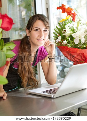 Side portrait of an attractive florist business woman owner at a flower shop counter using a laptop computer to place a stock order on line, smiling. Small business technology.