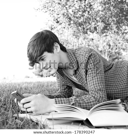 Black and white side portrait of a teenager student boy laying down in a park studying with his college books and using a smartphone to network, smiling. Technology lifestyle.