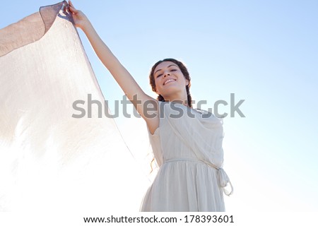 Beauty portrait view of an elegant smiling woman rising a floating silk fabric up with her arms against a bright blue sky on holiday, outdoors. Travel, beauty and lifestyle.