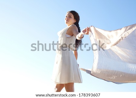 Side view of an elegant smiling woman rising a floating silk fabric up with her arms against a bright blue sky on holiday, outdoors. Travel, beauty and lifestyle.