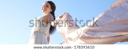 Panoramic side view of an elegant and aspirational smiling woman rising a floating silk fabric up with her arms against a bright blue sky on holiday, outdoors. Travel, beauty and lifestyle.