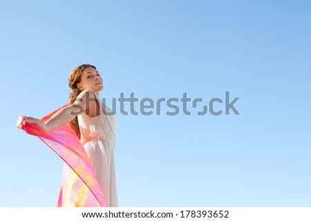Side beauty portrait of an elegant young woman rising a colorful floating silk fabric up with her arms against a bright blue sky on holiday, outdoors. Travel, beauty and lifestyle.
