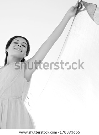 Black and white beauty portrait view of an elegant smiling woman rising a floating silk fabric up with her arms against a bright sunny sky on holiday, outdoors. Travel, beauty and lifestyle.