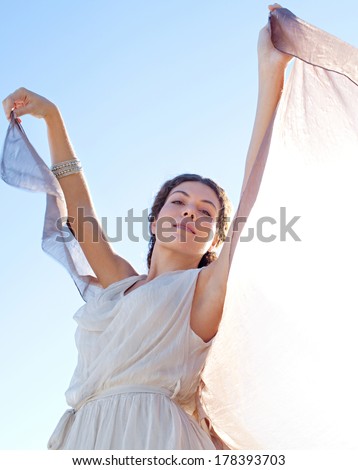 Beauty portrait close up of an elegant smiling woman rising a floating silk fabric up with her arms against a bright blue sky on holiday, outdoors. Travel, beauty and lifestyle.