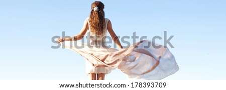 Panoramic rear view of a young woman holding a floating silk fabric with her hands against a bright blue sky on a holiday, travel outdoors. Beauty and healthy lifestyle.