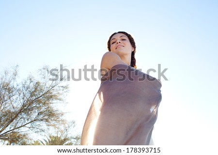 Beauty portrait view of an elegant woman wrapping her body in floating silk fabric, joyfully smiling against a bright blue sky on holiday, outdoors. Travel, beauty and lifestyle.