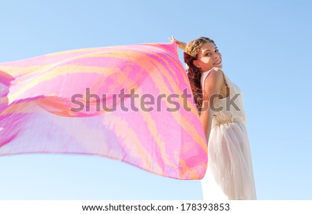 Side beauty portrait of an elegant young woman rising a colorful floating silk fabric up against a bright blue sky, joyfully having fun on holiday, outdoors. Travel, beauty and lifestyle.