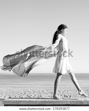 Black and white side view of an elegant woman rising a floating silk fabric up with her arms against a sunny sky, traveling on a holiday beach. Taking a step on a stone wall, beauty and lifestyle.