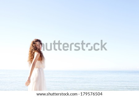 Attractive young woman relaxing on a blue sea and sky beach wearing a white silk dress and turning to camera thoughtfully against a blue sky. Outdoors healthy and well being lifestyle.