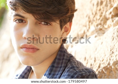 Close up portrait of an attractive teenager student boy leaning on an old and aged stone wall in a park during a sunny day, looking at the camera with a thoughtful expression, outdoors.