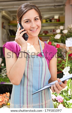 Joyful florist business woman working at her flower market store and using the phone to have a conversation and order stock. Small business owner running a retail shop, outdoors.