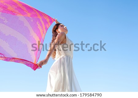 Side view of a beautiful young woman holding a pink silk fabric up in the air floating with the breeze against a vivid blue sky during a summer holiday. Outdoors wellness lifestyle.