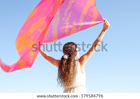 Rear view of a young woman with her arms up in the air holding a pink silk fabric floating with the breeze during a sunny summer day on holiday, against a bright blue sky. Outdoors beauty lifestyle.