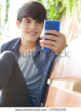Close up portrait of a beautiful teenager boy sitting and relaxing on a wooden park bench, holding and using his smartphone device to network and browse on-line. Technology lifestyle.