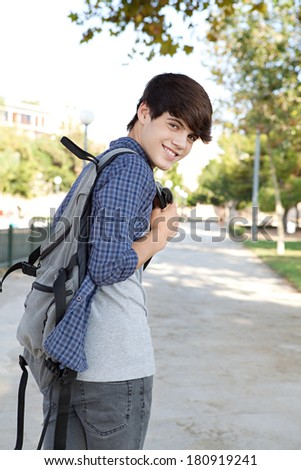 Rear view of a teenager student boy walking on a path at a college campus grounds carrying a backpack and turning to smile at the camera joyfully. Student lifestyle outdoors.