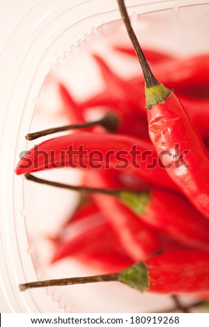 Over head close up view of a bunch of spicy red hot chili peppers together in a transparent plastic container against a white background. Hot food ingredient, indoors.
