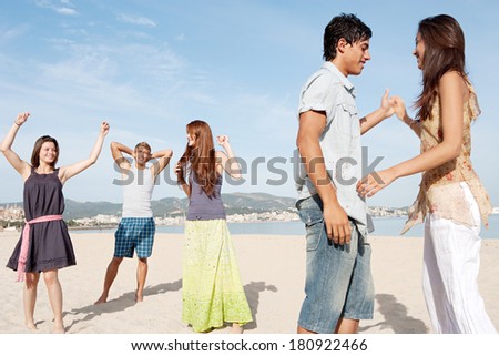Group of five teenagers friends enjoying a day on the beach together, listening to music and dancing, celebrating the summer holidays during a sunny day with a blue sky.