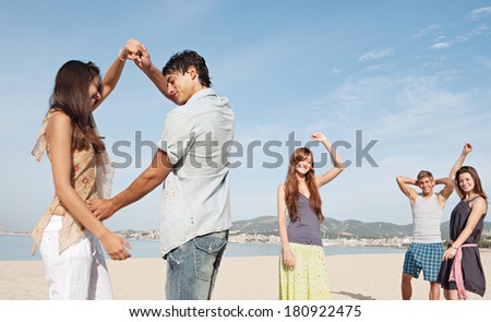 Group of five teenagers tourists friends enjoying a day on the beach together, listening to music and dancing, celebrating the summer holidays during a sunny day with a blue sky.
