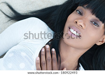 Over head close up beauty portrait of a young and attractive african american black woman with perfect skin on a beach, laying down on sand and softly smiling. Health and beauty outdoors lifestyle.