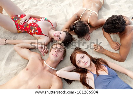 Over head portrait of a group of five teenagers boys and girls laying down together on a golden sand beach, relaxing during a sunny day on holiday. Travel and healthy lifestyle.