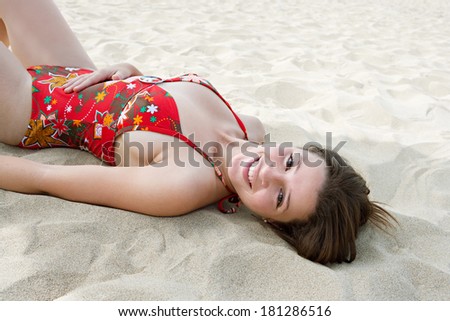 Side portrait view of a beautiful young woman laying down on a golden sand beach on holiday enjoying a summer day sunbathing, turning to smile at the camera. Travel and beauty lifestyle.