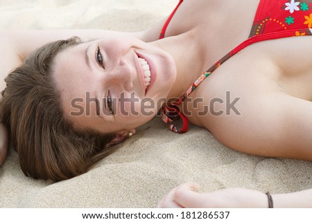 Over head close up portrait of a beautiful young woman laying down on a golden sand beach on holiday enjoying a summer day sunbathing, turning to smile at the camera. Travel and beauty lifestyle.
