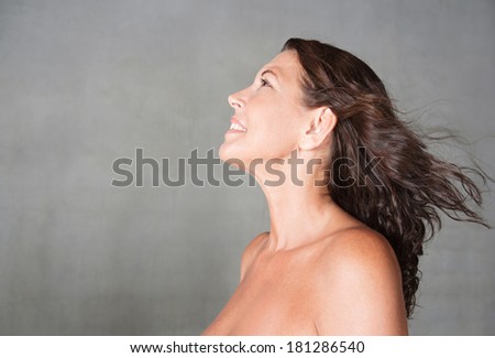 Close up profile portrait of a beautiful mature woman looking up standing against a plain gray wall with her hair flying in the blowing breeze, feeling fresh and healthy. Beauty and care lifestyle.
