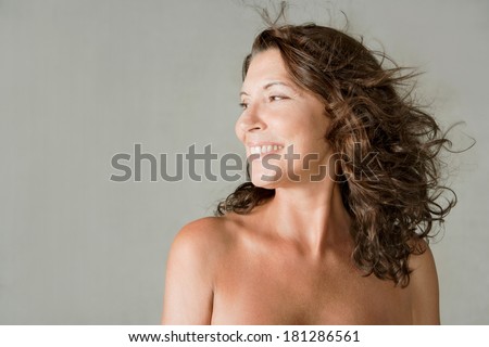 Close up beauty portrait of an elegant mature woman with bare shoulders, smiling against a plain gray wall background while her hair is flying and floating in the breeze. Beauty lifestyle.