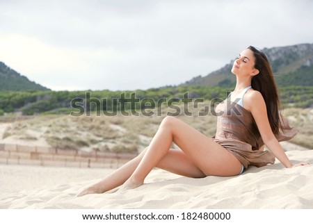 Close up of attractive young woman with perfect legs and skin relaxing on the sand dunes of a beach with mountains, in a silk fabric sarong with her hair blowing in the breeze. Health and lifestyle.