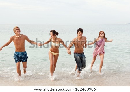 Energetic group of four friends laughing running together on the shore of a white sand beach on holiday, joyfully having fun and enjoying an exciting trip. Travel and teenagers lifestyle.