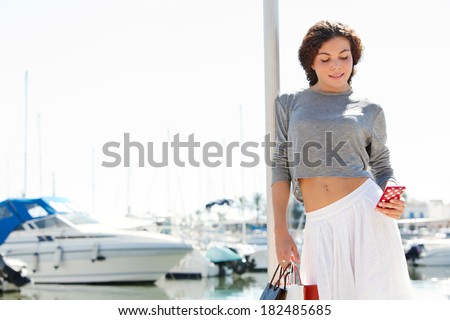 Beautiful young woman relaxing in a boats and yachts marine port carrying paper shopping bags and using on a smartphone during a sunny day on holiday. Travel technology lifestyle.