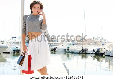 Beautiful young woman relaxing in a boats and yachts marine port carrying paper shopping bags and calling on a smartphone during a sunny day on vacation. Travel technology lifestyle.