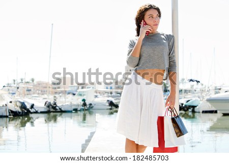 Beautiful young woman relaxing in a boats and yachts marine port carrying shopping bags and calling on a smartphone during a sunny day on vacation. Travel technology lifestyle.
