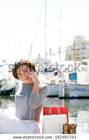 Attractive young woman sitting down and relaxing in a yachts port marina with shopping carrier bags and using a smartphone to make a call during a sunny day on vacation. Technology lifestyle.
