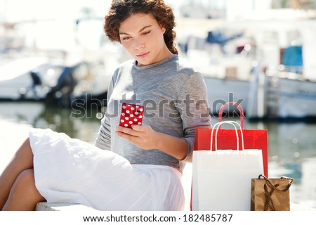 Attractive young woman sitting down and relaxing in a yachts port marina with shopping carrier bags and using a smartphone during a sunny day on vacation. Travel technology lifestyle.