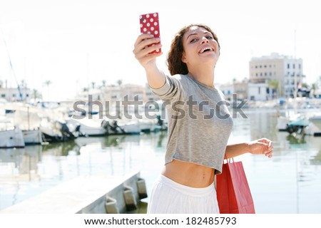 Active and attractive young woman having fun on holiday visiting a coastal town with a yachts marine port during a sunny day, taking a selfie photo of herself. Technology and travel lifestyle.