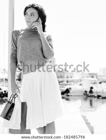 Black and white portrait of a young woman relaxing in a boats port carrying shopping bags and calling on a smartphone during a sunny day on vacation. Travel technology lifestyle.