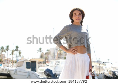 Attractive young woman walking in a boats and yachts marine pier carrying paper shopping bags and smiling joyfully at the camera during a sunny day on holiday. Travel lifestyle.