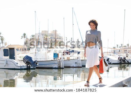 Beautiful young woman walking in a boats and yachts marine pier carrying paper shopping bags and smiling joyfully at the camera during a sunny day on holiday. Travel lifestyle.