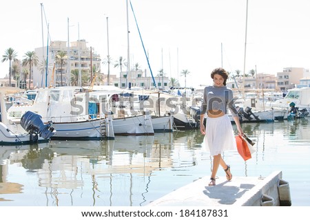 Attractive young woman walking in a boats and yachts marine pier carrying paper shopping bags and smiling joyfully during a sunny day on holiday. Travel lifestyle.