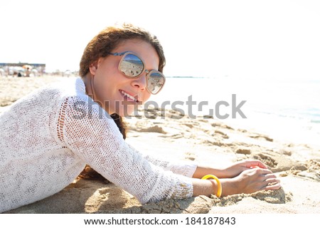 Portrait of an attractive young woman laying down on a golden sand beach on holiday smiling at the camera and wearing protective shades. Travel and healthy lifestyle, outdoors.